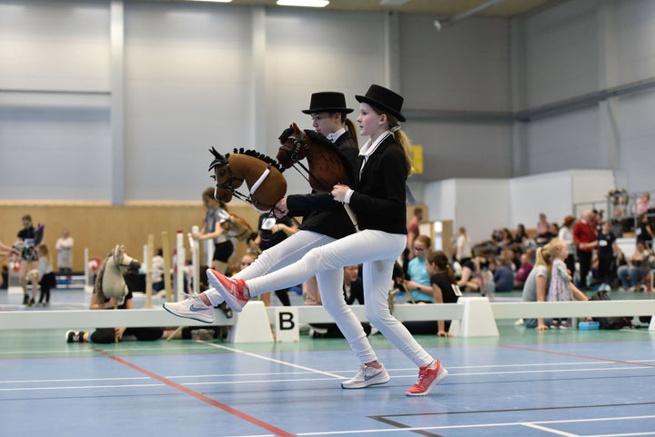 Två barn i formell tävlingsklädsel för Hobby Horsing tävlar inomhus med sina käpphästar