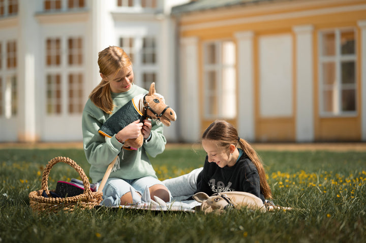 En flicka i ljusgrön sweatshirt sitter i gräset bredvid en flätad korg med utrustning för käpphästar och håller varsamt en ljusbrun käpphäst med svart träns, vit man och quiltat täcke. Ett annat barn syns delvis på en filt i närheten, i en lugn stämning.