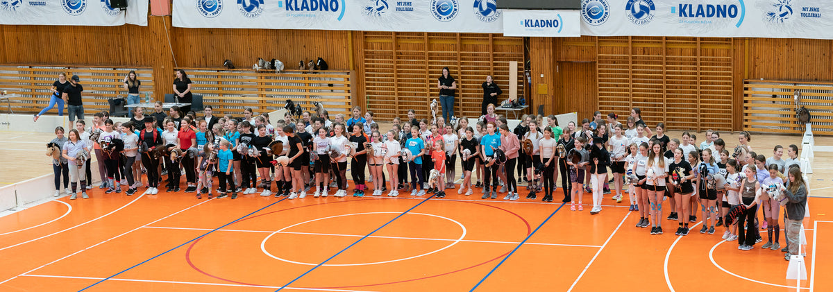 En stor grupp unga tävlande i Hobby Horsing står på rad över en orange idrottsplan inomhus i sportiga fritidskläder och håller många olika käpphästar, med ribbstolar i trä, läktare och banderoller från Kladno i bakgrunden.
