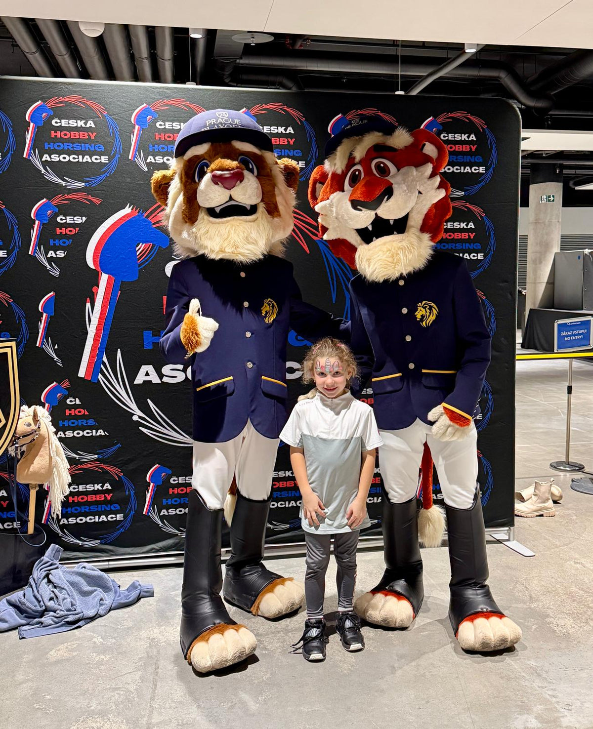 A smiling young girl with face paint stands between two Prague Playoffs animal mascots in navy riding-style jackets and caps, posing in front of a Česká Hobby Horsing Asociace backdrop in the O2 Arena concourse. A small hobby horse rests at the side