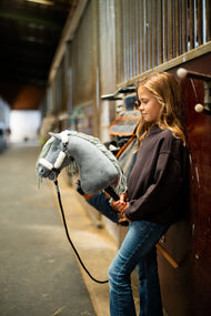 Girl in a dark sweatshirt in a stable aisle with Hobby Horse Calypso in a fur-lined halter and black lead rope