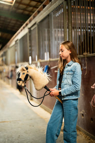 Girl in a denim jacket and jeans holds a cream Hobby Horse with black fur halter on the black reins in a stable aisle