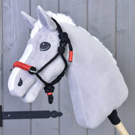 Grey plush Hobby Horse with a white mane wearing the black and red rope halter in front of a grey wooden wall
