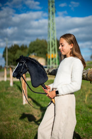 Flicka i vit fluffig tröja på en solig äng som håller Hobby Horse Loki i svart träns med tyglar