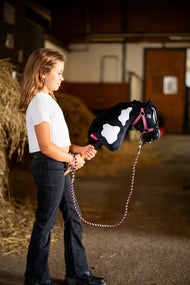 Girl in a white top stands in a stable with Hobby Horse Merlin with Black Halter, here on a pink halter and braided lead rope