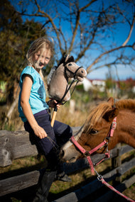 Leende blond flicka på ett trästaket med en beige käpphäst i svart träns, bredvid en fuxfärgad ponny i rosa grimma