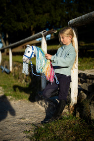 En flicka i mintgrön sweatshirt vid ett vitt staket på en ridbana håller en vit käpphäst med man i regnbågens färger i det blårandiga grimskaftet