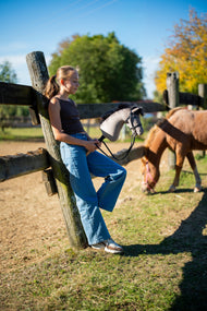 Flicka lutad mot ett hagstaket i höstsol med Hobby Horse Thor i svart träns, en fuxfärgad ponny som betar bakom