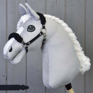 Grey plush Hobby Horse with a braided white mane wearing the black halter with black fur in front of a grey wooden wall