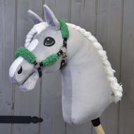 Grey plush Hobby Horse with a braided white mane wearing the black halter with green fur in front of a grey wooden wall