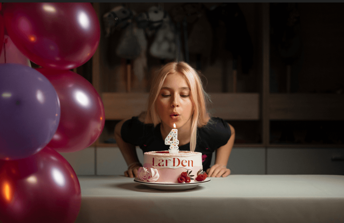 Lara blåser ut ett ljus format som en fyra på en födelsedagstårta från LarDen bredvid färgglada ballonger