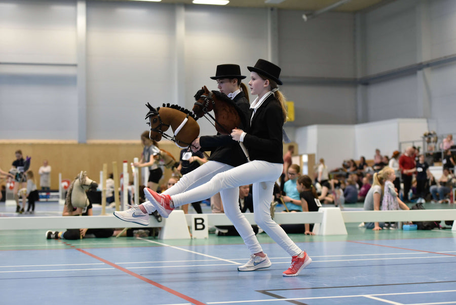 Two children in formal Hobby Horsing dress code outfits competing indoors with Hobby Horses