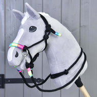 Grey Hobby Horse with white blaze wearing the black rope halter and reins with rainbow wraps by a grey wooden wall