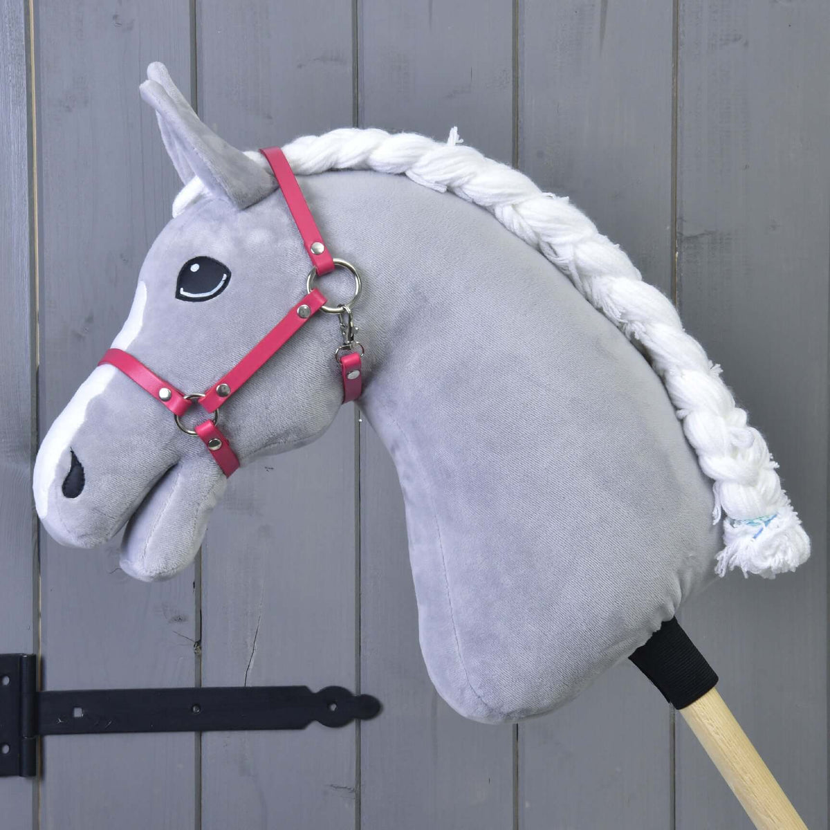 A grey plush Hobby Horse with a white blaze and braided mane wears the raspberry pink halter by a grey wooden wall