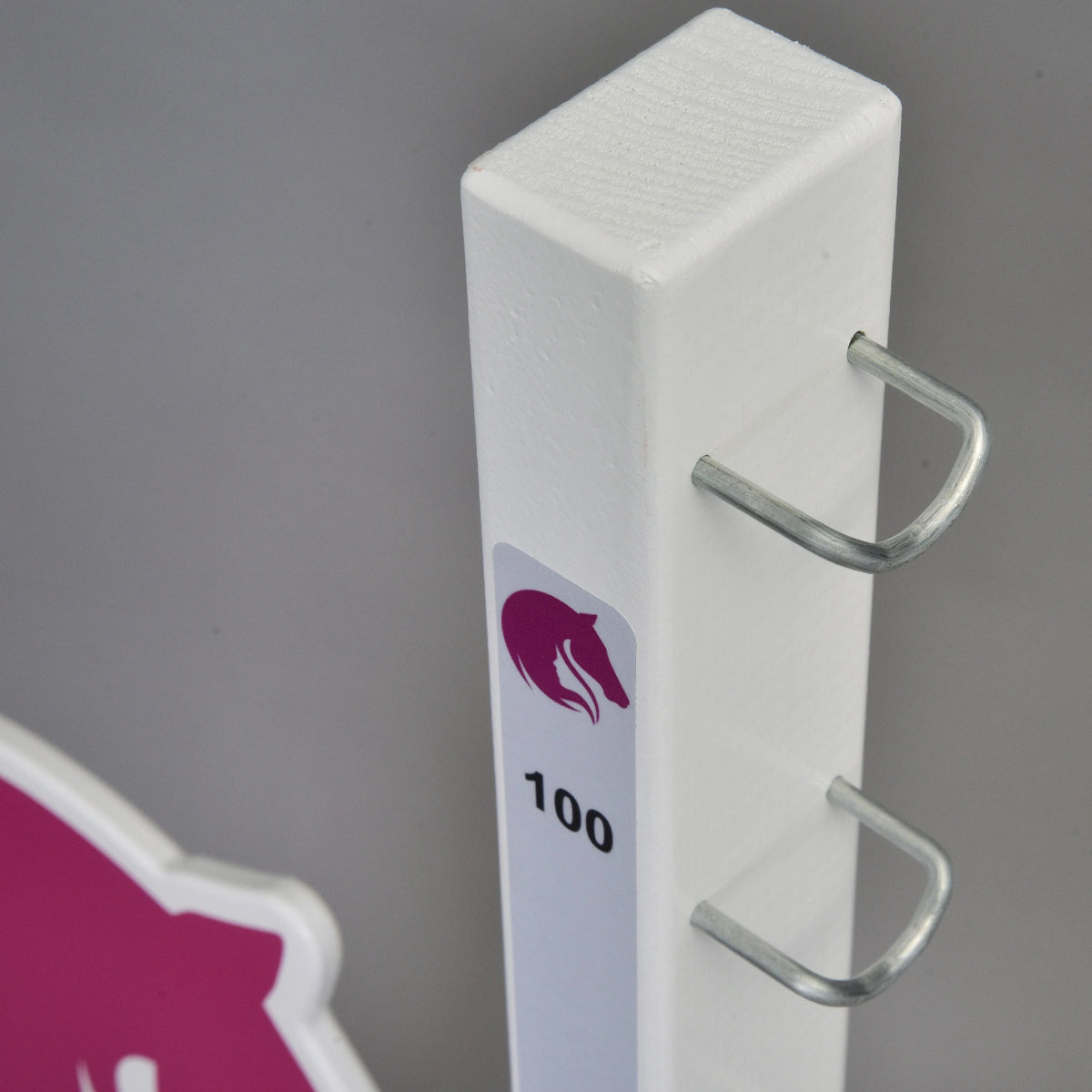 Närbild av toppen av en vit stolpe med U-formade metallkrokar och en liten rosa hästlogotyp ovanför markeringen 100