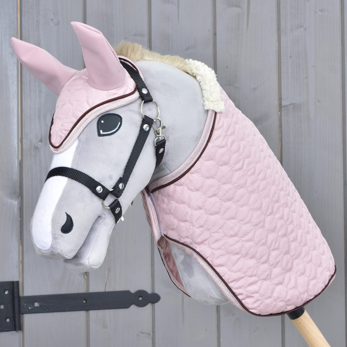 Grey Hobby Horse in a black halter wearing the Set Blush, a pink quilted blanket and ear bonnet, against a grey wooden wall