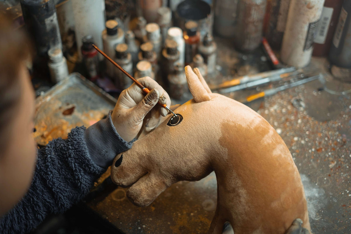 Craftsperson painting the eye of a light brown plush Hobby Horse head at a workbench full of paint bottles