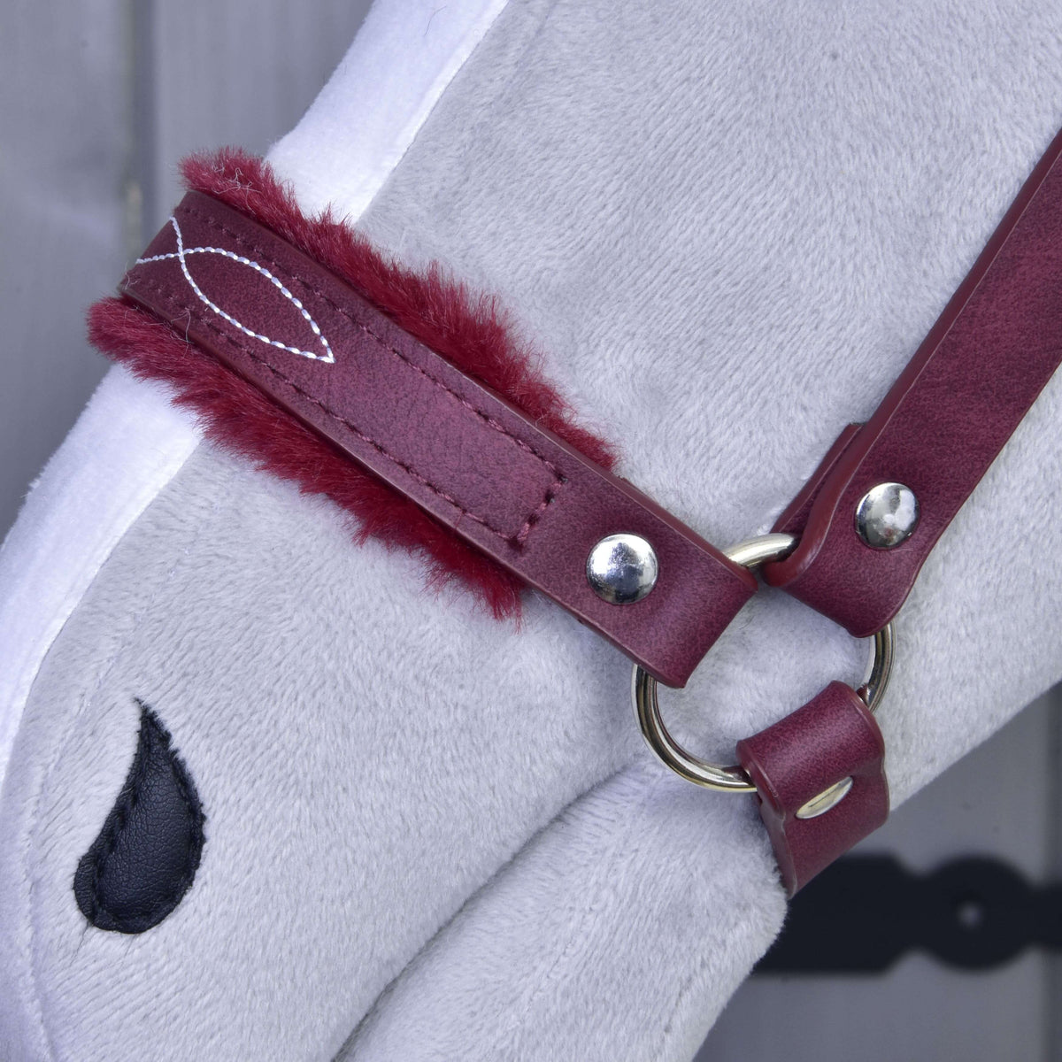 Close-up of the burgundy noseband with fur trim, white decorative stitching and a silver ring on a grey Hobby Horse