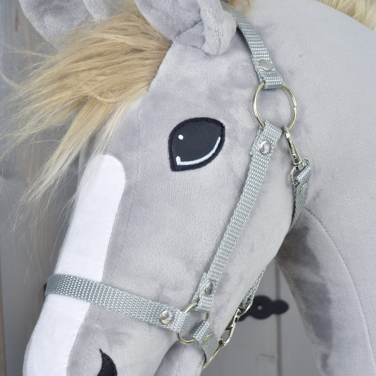 Close-up of the grey webbing halter with silver rings and rivets around the embroidered eye of a grey plush head