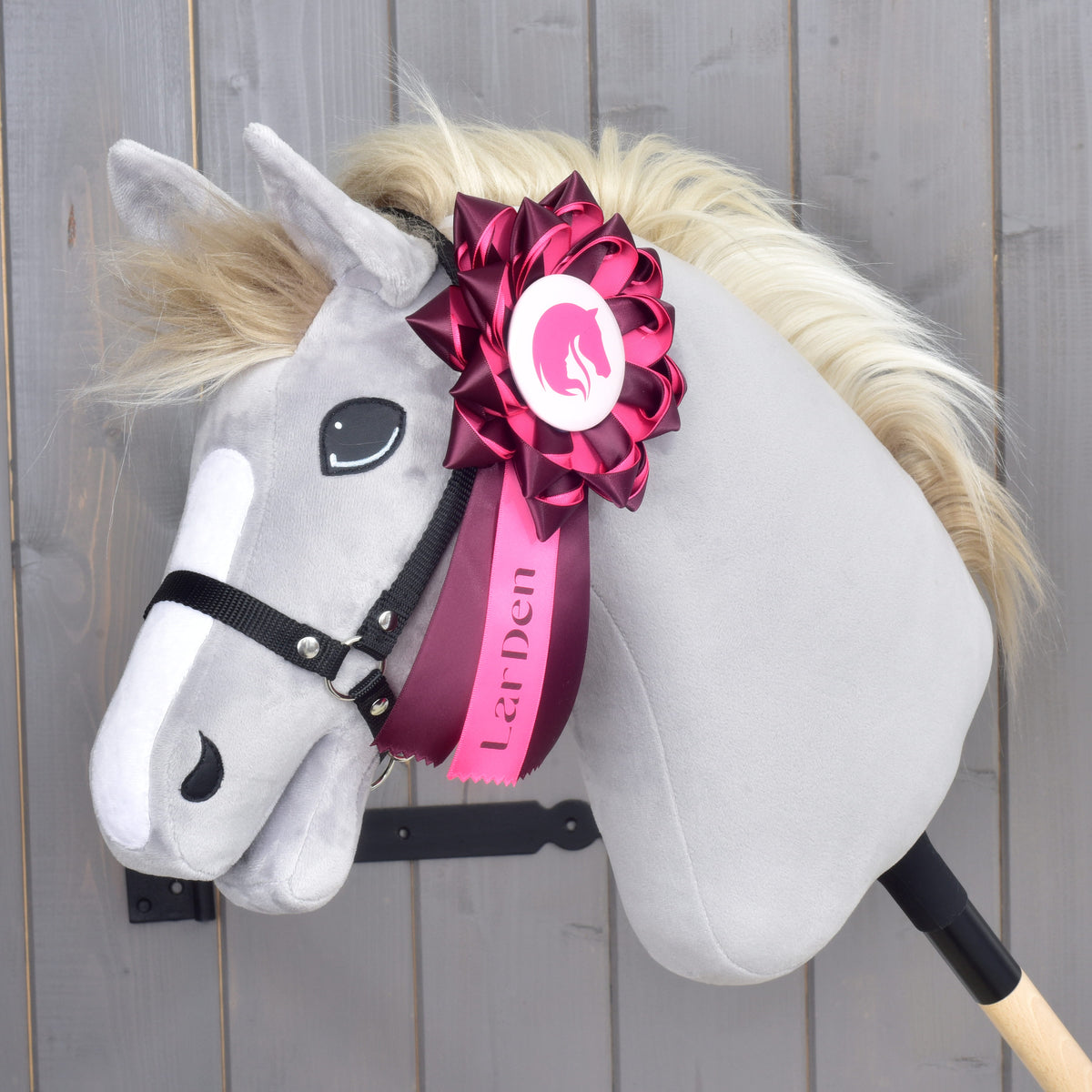 Hobby Horse rosett Raspberry Rose på den svarta grimman på ett grått plyschhuvud, med texten LarDen tryckt på det rosa mittbandet