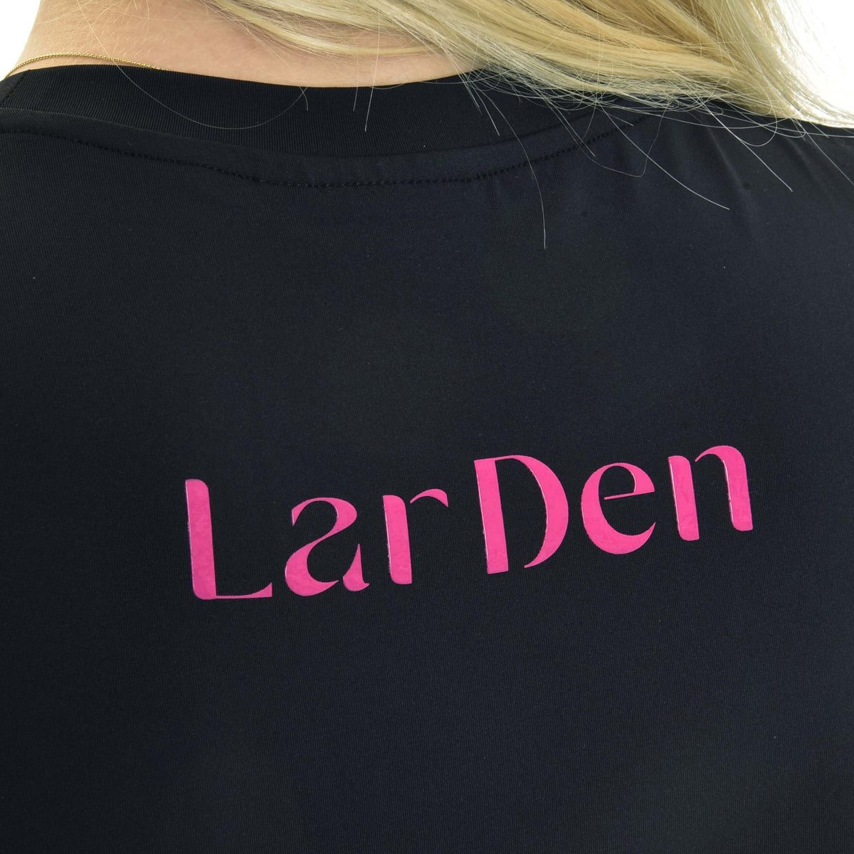Närbild av den rosa LarDen-texten på ryggen av en svart sporttröja med rund hals, blont hår över axeln
