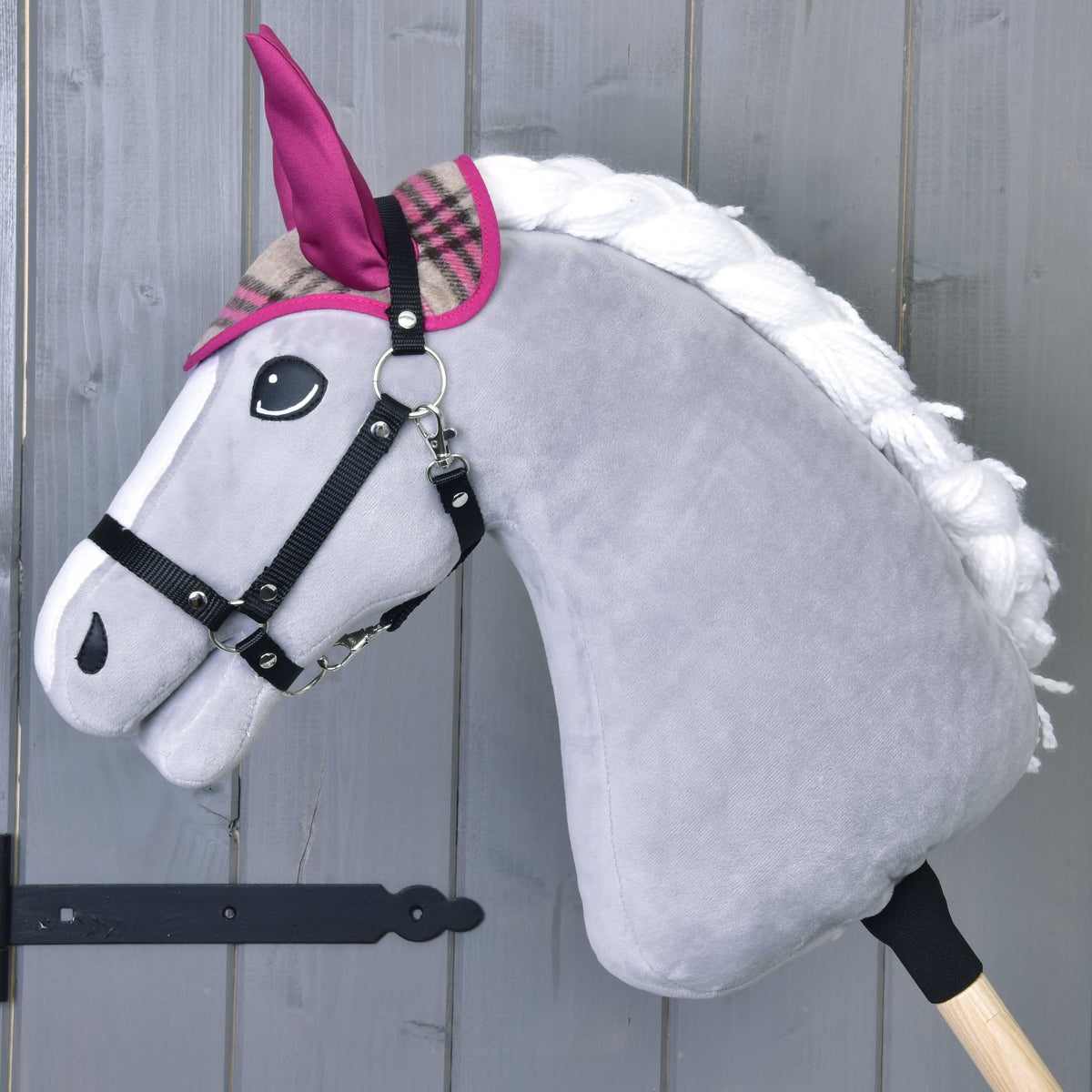 Grå käpphäst i svart grimma som bara bär den rutiga flughuvan med rosa öron och rosa kantband