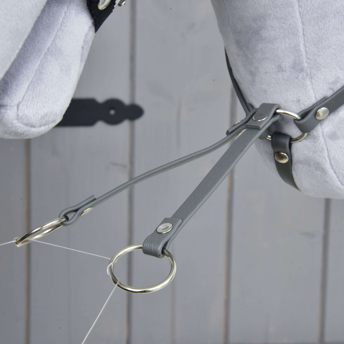 Närbild av de två gaffelremmarna på den mörkgrå martingalen av konstläder som slutar i silverfärgade ringar under plyschhuvudet