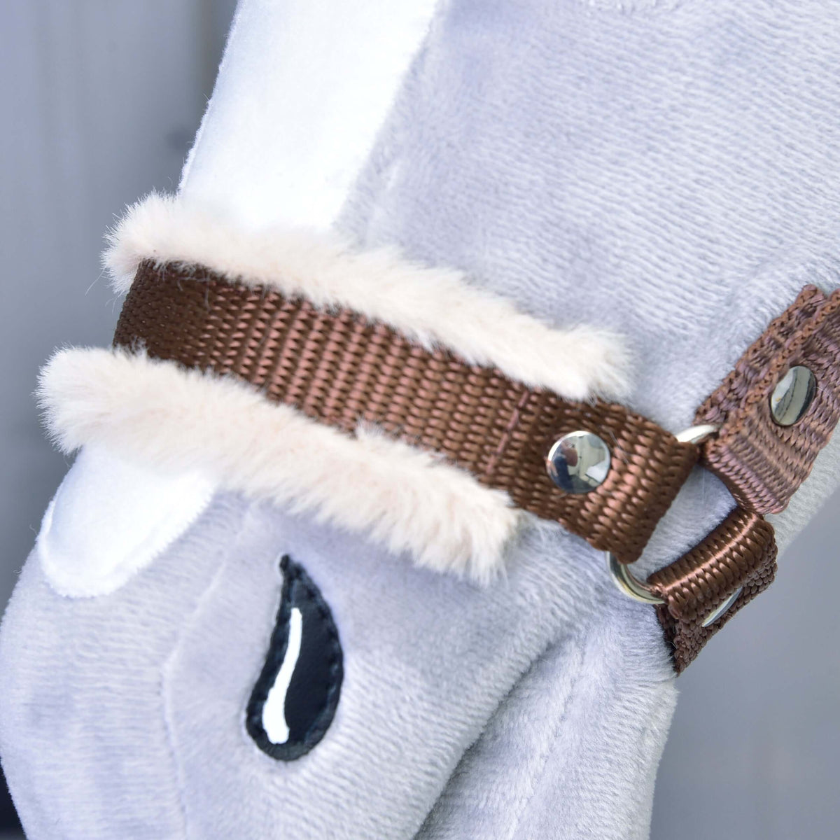 Närbild på den bruna nosremmen av vävt band med mjuk beige päls och en silvernit på en grå käpphäst i plysch