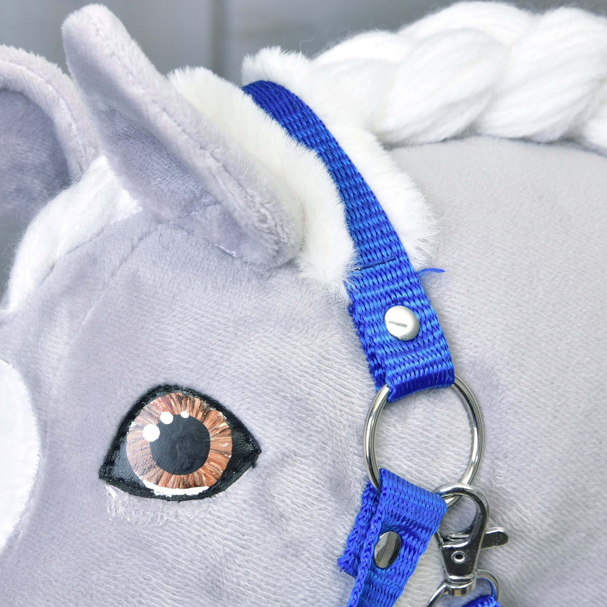 Close-up of the blue crownpiece with white fur behind the ear of a grey Hobby Horse, with silver ring and snap hook