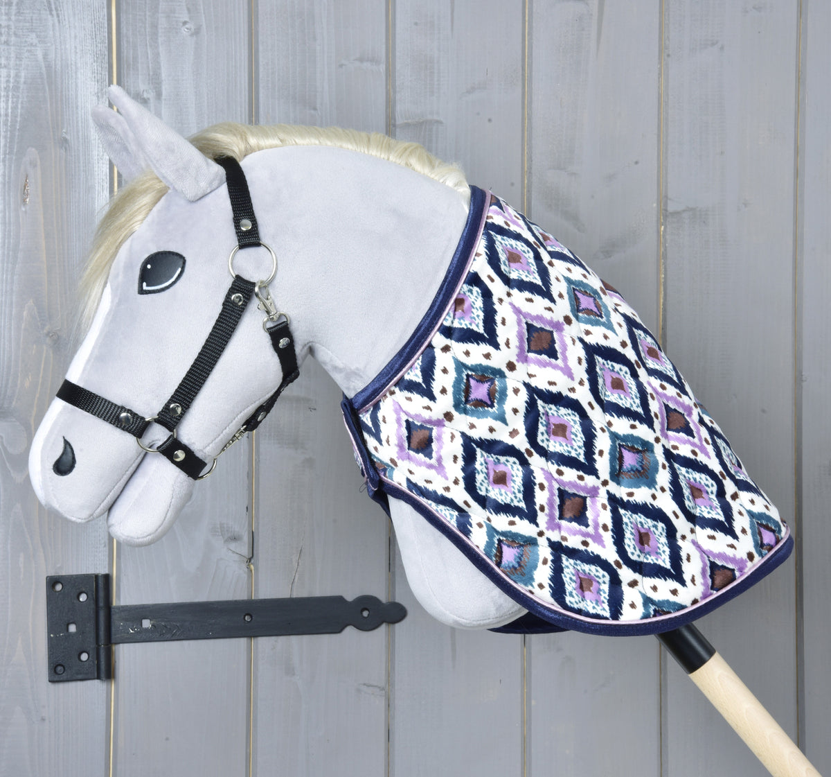 Grey Hobby Horse with a black halter in left profile, the Indian blanket covering its neck, grey wooden wall behind