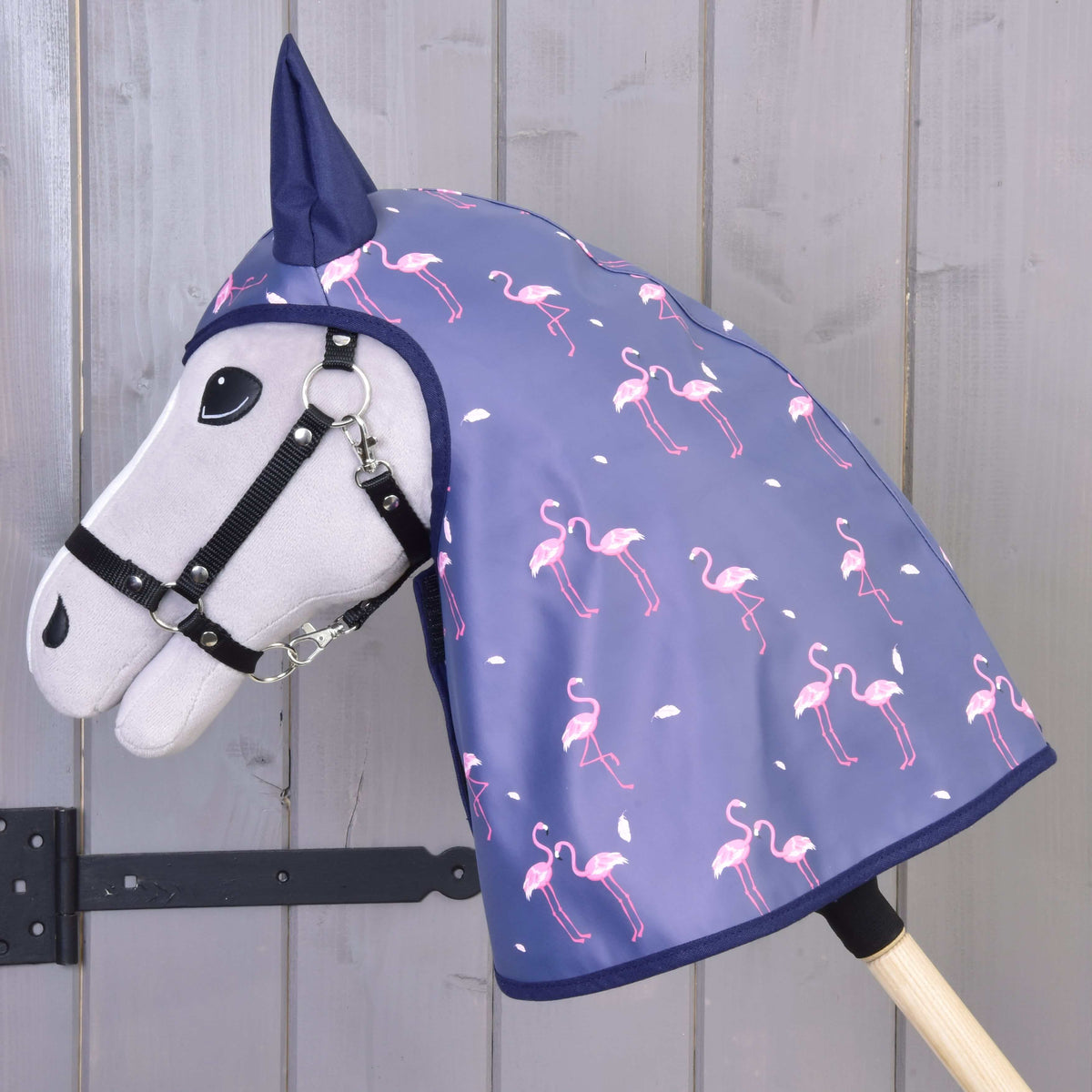 Side view of the flamingo print raincoat covering the head and neck of a grey Hobby Horse by a grey wooden wall