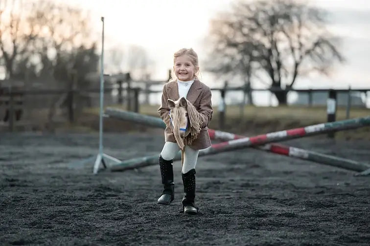 Liten flicka i tweedjacka och ridstövlar som leker med en krämvit käpphäst på en sandbana med bommar
