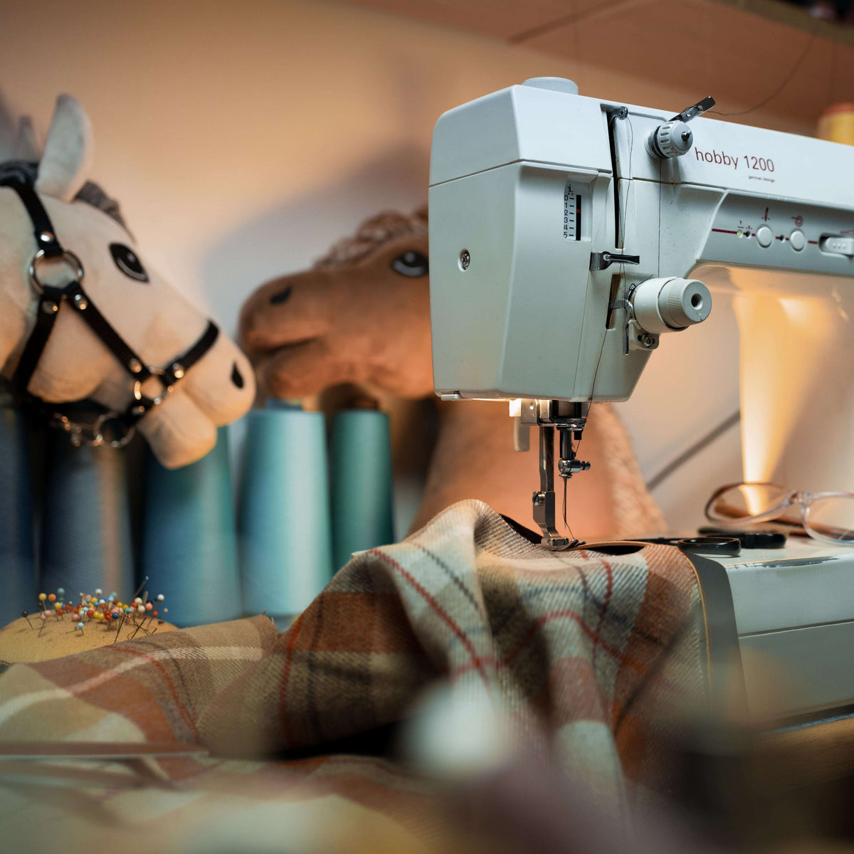 Workshop corner: a sewing machine stitches plaid wool while a cream Hobby Horse in a black halter looks on