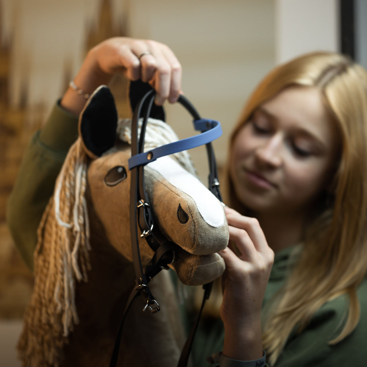 Blonde girl fitting a dark brown bridle with a blue browband onto a light brown Hobby Horse with a blonde yarn mane