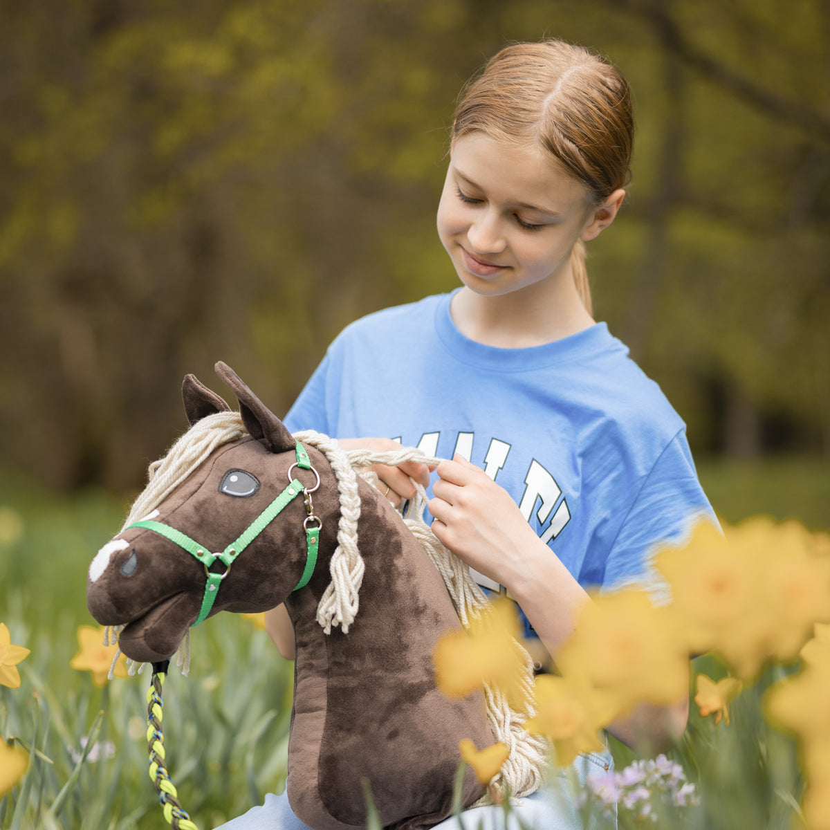 Flicka i blå T-shirt som flätar manen på en mörkbrun käpphäst i grön grimma bland gula påskliljor