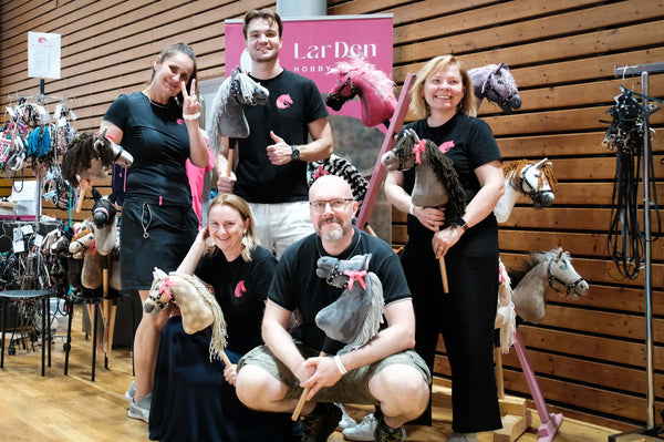Five smiling people in black T-shirts posing with Hobby Horses at a stand with a pink LarDen banner