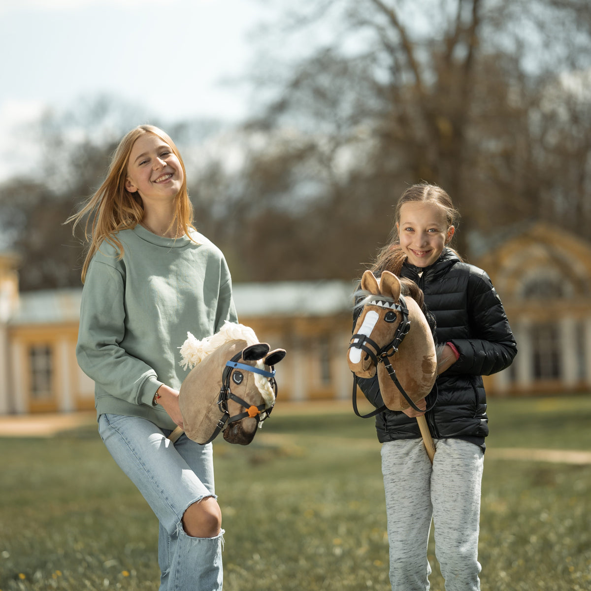 Two smiling girls in a park with a beige and a light brown Hobby Horse in black bridles, a yellow building behind