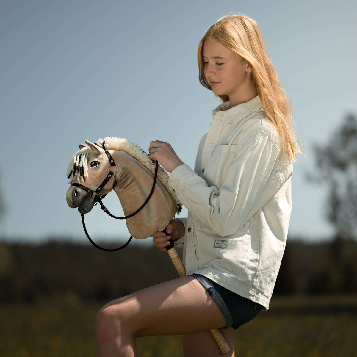 Blond flicka i krämvit jeansjacka som håller en black käpphäst med svart träns och tyglar mot en blå himmel