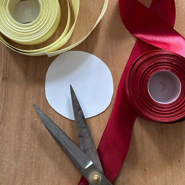 Rolls of pale yellow and red satin ribbon, a cut-out paper circle and old scissors on a wooden table