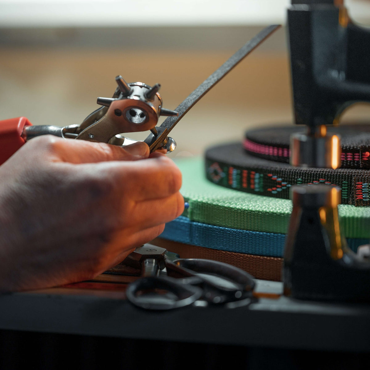 Workbench close-up: punching holes in a black strap, with rolls of coloured webbing and scissors in the background