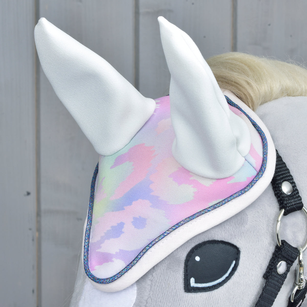 Närbild på Hobby Horse-flughuvan med vita öron, pastellfärgat leopardmönster och glittrande blå kantband på en grå häst