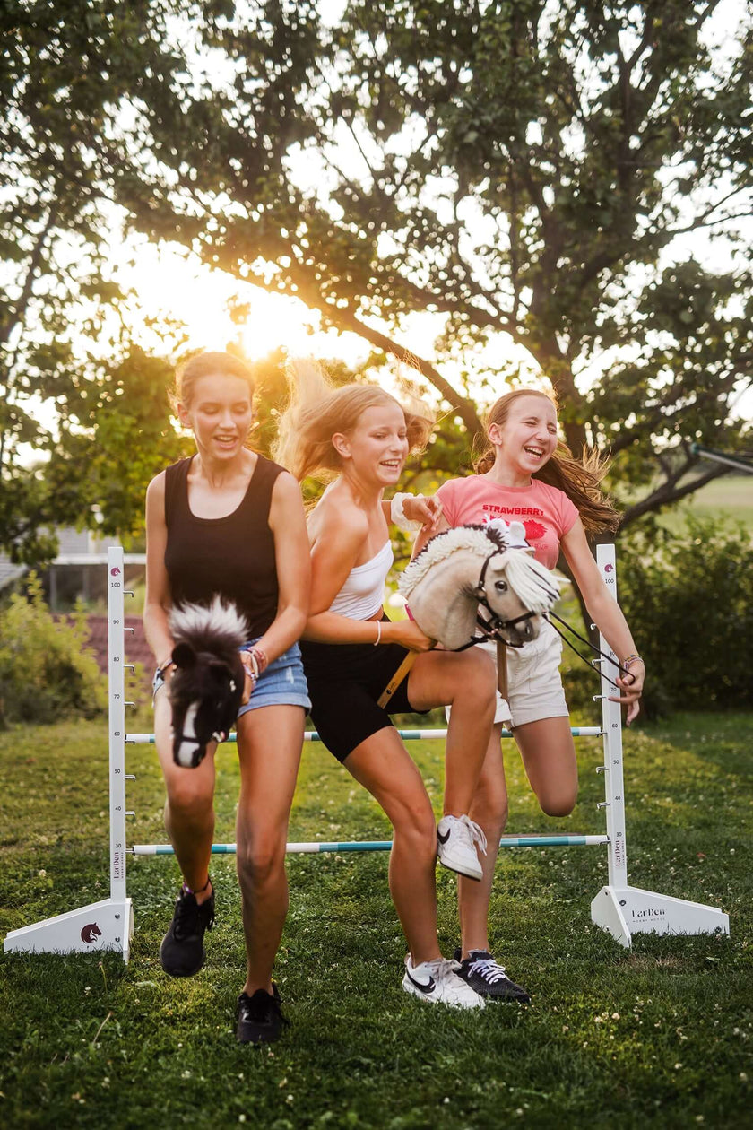 Three laughing girls jumping a LarDen jump with their Hobby Horses in a garden at sunset