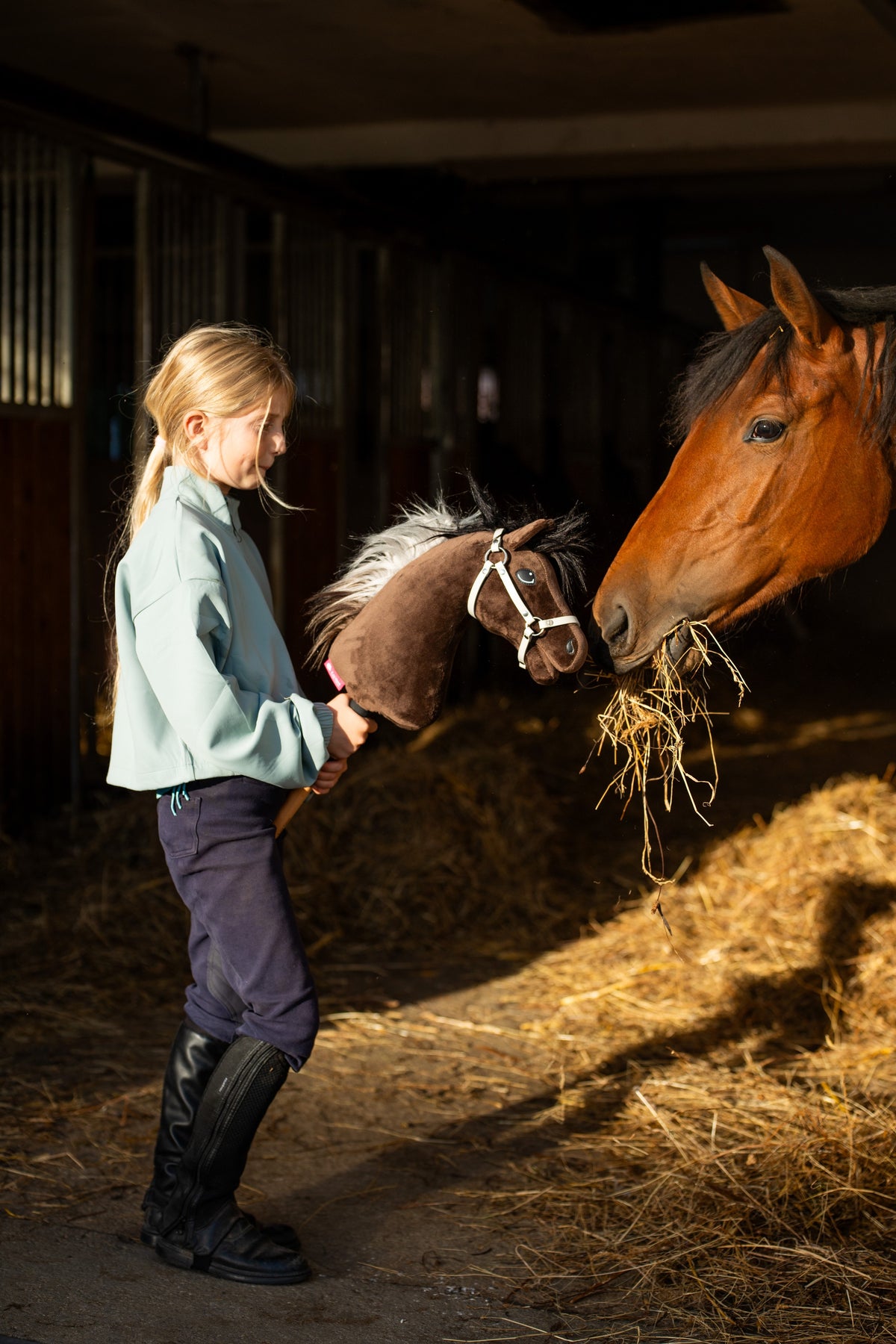 Flicka i mintgrön topp och ridstövlar i ett stall håller Hobby Horse Alfredo i vit grimma bredvid en brun häst som äter hö