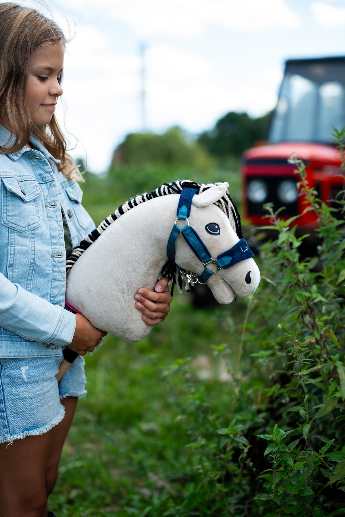 Girl in a denim jacket holding Hobby Horse Aurora in a blue halter outdoors by green plants, a red tractor behind
