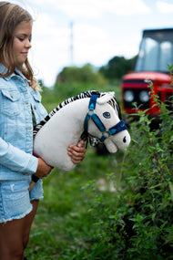 Flicka i jeansjacka som håller Hobby Horse Aurora i blå grimma utomhus vid gröna växter, med en röd traktor bakom