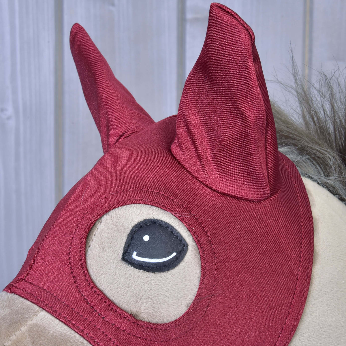 Detalj av de glansiga mörkröda öronskydden på masken Barn Red, med den sydda ögonöppningen nedanför, på en beige käpphäst