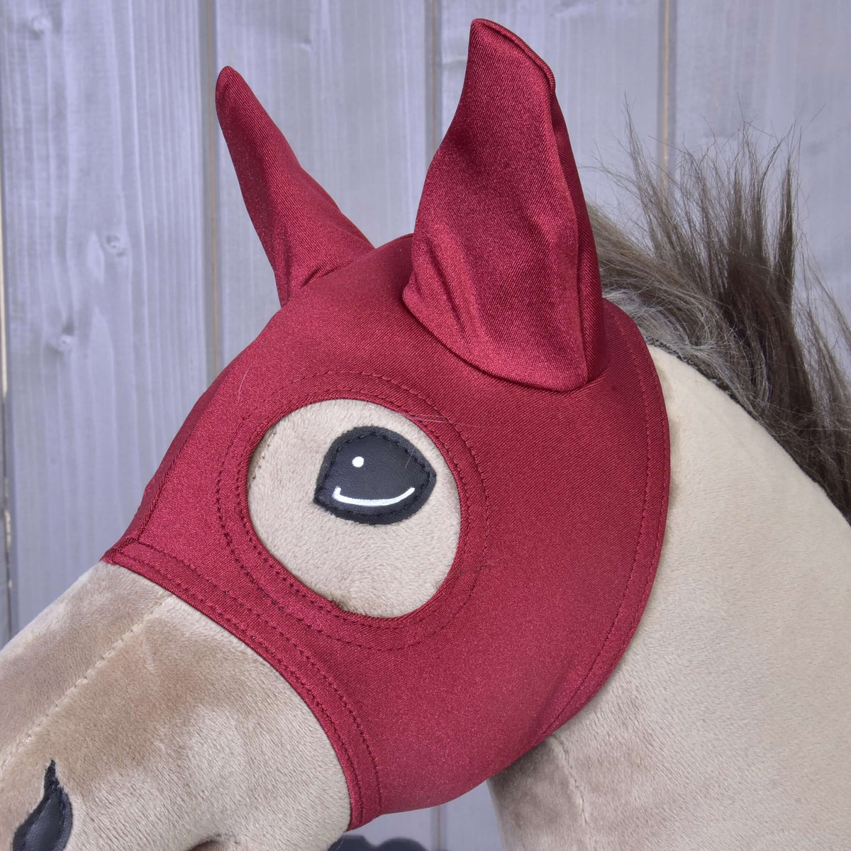 Närbild på den mörkröda masken Barn Red på en beige käpphäst, båda öronen i spetsiga skydd och ögat inramat av sömmar