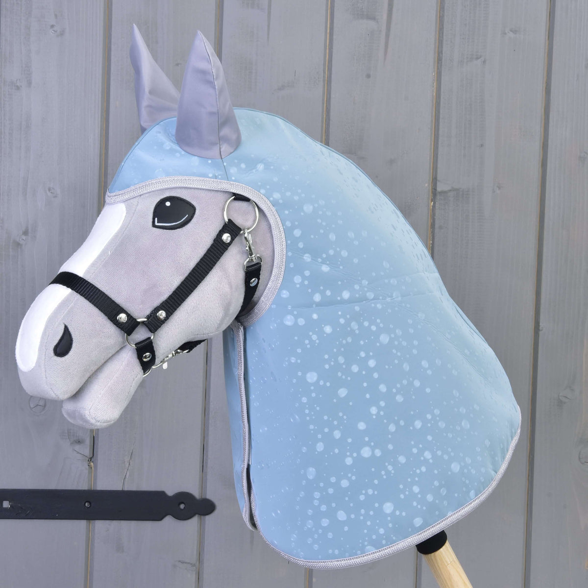 Regntäcke Blue Drops på en grå käpphäst med vit bläs och svart grimma, täcker huvud och hals, vid en grå plankvägg