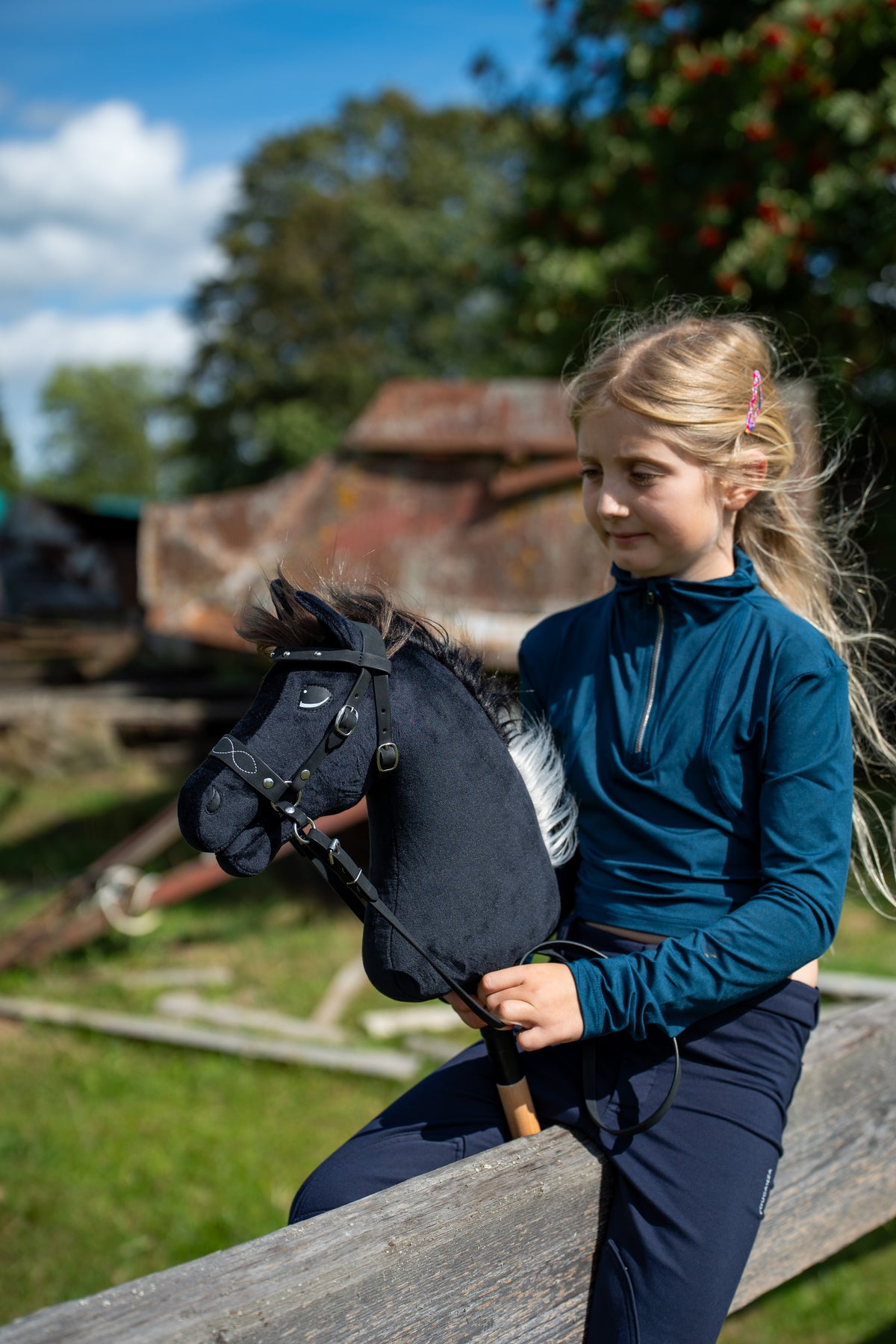 Blonde girl in a teal top sits on a wooden rail holding Hobby Horse Bond with Black Halter, here in a black bridle with reins