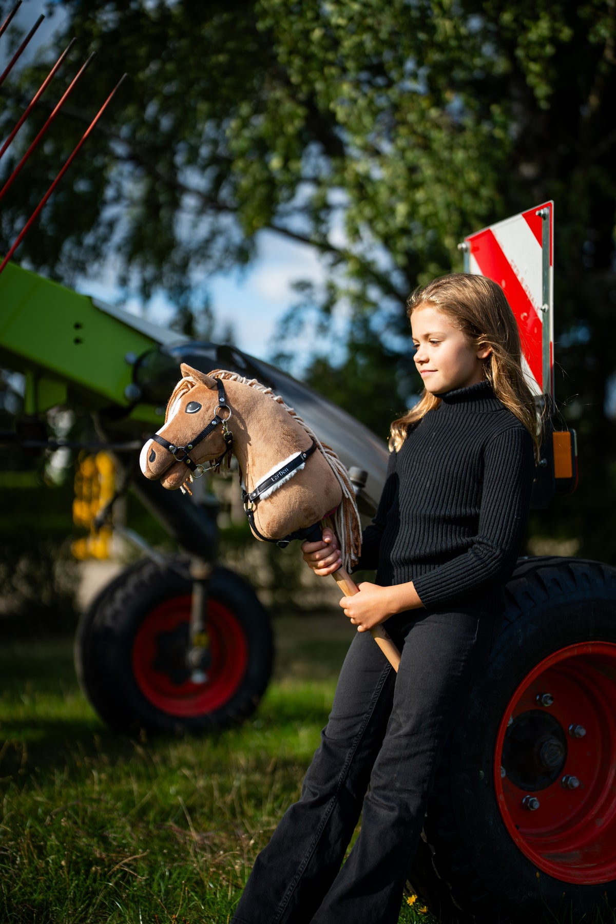 Flicka i svart polotröja vid gröna lantbruksmaskiner håller Hobby Horse Bren med svart grimma, här med svart träns